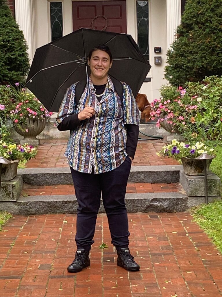 Jess stands on a red brick walkway that leads to a home. They're wearing black pants, and a multi colored vertical striped shirt over a black long sleeve shirt. They're smiling as they hold a black umbrella over their head. Jess Falero on the campaign trail.