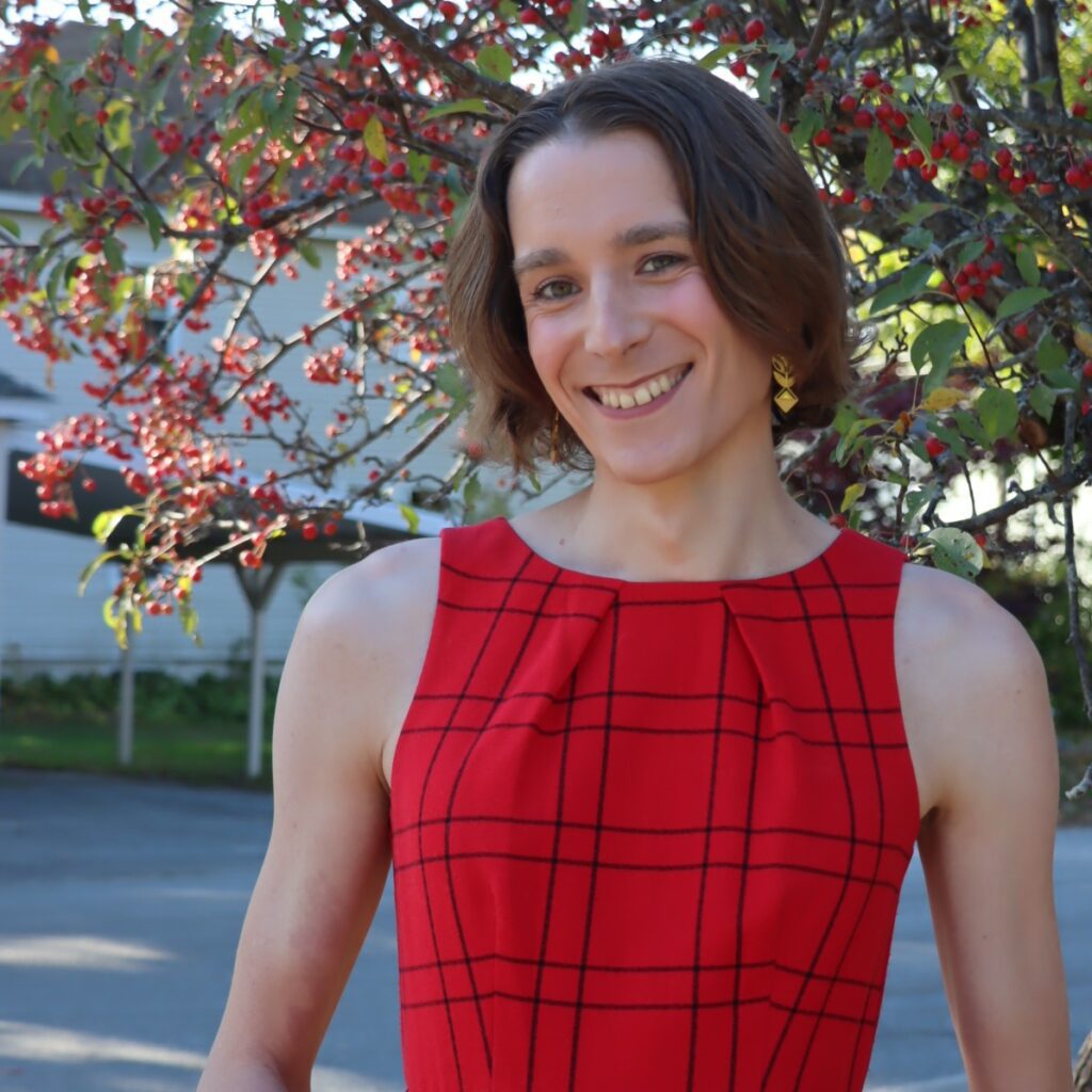 Ramona Thieme poses with a smile in front of some branches of a flower tree. She is wearing a short sleeve dress that is red with a black plaid pattern on it.