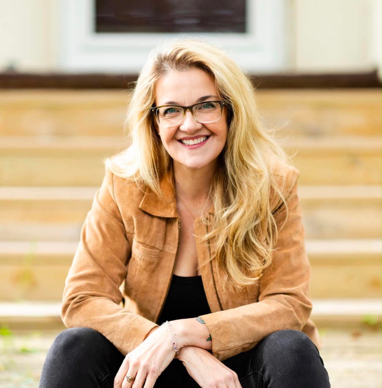 Amy is seated on some stairs and smiles at the camera. Her hair is long and blond. She wears glasses, black pants and top, with a light brown jacket on top. Her arms are propped on her knees and she crosses them at the wrist.