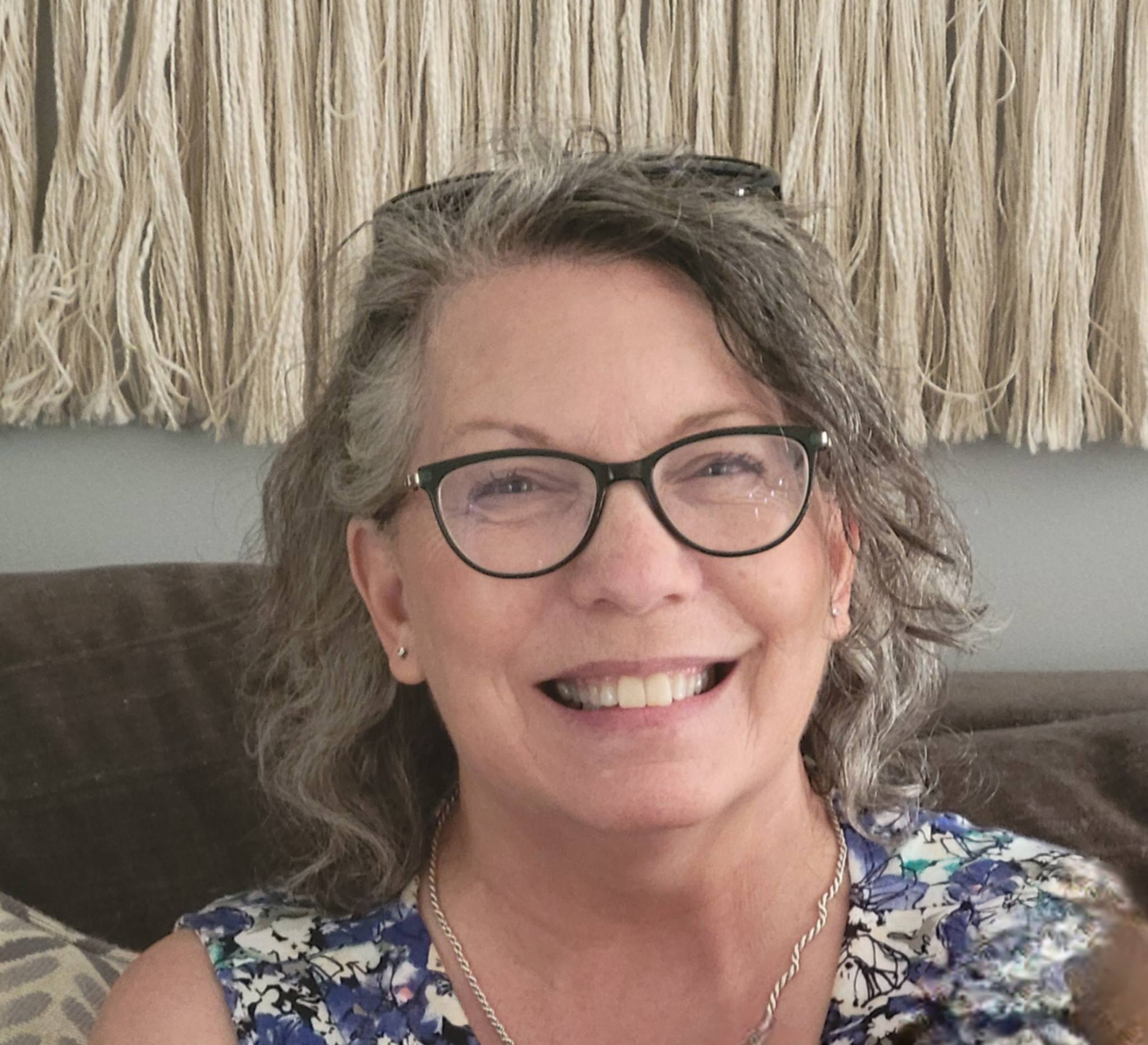 April smiles at the camera. She is wearing black rimmed glasses, has light grey and dark grey hair that is wavy and just reaches her shoulders. She is wearing a floral printed dress.
